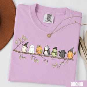 Peut inclure: Un t-shirt violet clair orn&eacute; d'un motif repr&eacute;sentant sept petits oiseaux perch&eacute;s sur une branche, chacun portant un costume d'Halloween diff&eacute;rent, notamment un chapeau de sorci&egrave;re, un drap de fant&ocirc;me, une citrouille, une cape de vampire et des ailes.