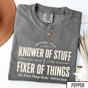 May include: A gray Comfort Colors t-shirt with the text "OFFICIAL TITLE KNOWER OF STUFF FIXER OF THINGS". The shirt has a crew neck and short sleeves. A silver necklace with a pendant is draped across the shirt.