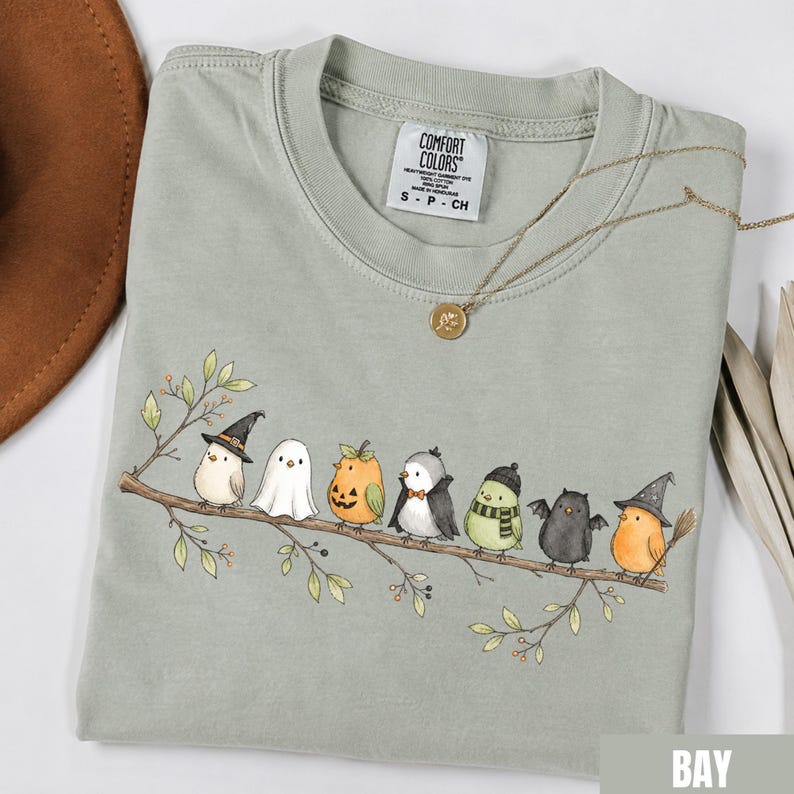 Peut inclure: Un t-shirt &agrave; col rond vert sauge avec une illustration de sept petits oiseaux perch&eacute;s sur une branche, chacun portant un costume d'Halloween diff&eacute;rent, notamment un chapeau de sorci&egrave;re, un drap de fant&ocirc;me, une citrouille et des ailes de chauve-souris.
