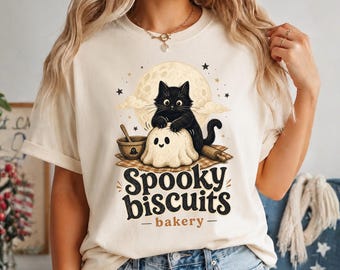 May include: A cream-colored t-shirt featuring a graphic of a black cat kneading a ghost-shaped dough on a checkered cloth. The design includes a full moon, a mixing bowl, a rolling pin, and the text Spooky biscuits bakery in black and brown.