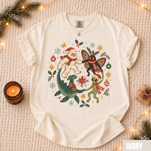 Peut inclure: Un t-shirt ivoire avec un motif circulaire représentant un yéti, un homme-phalène, un monstre du loch ness et un extraterrestre, portant des écharpes rayées rouge et blanc et des bonnets de Noël, entourés de boules, de houx et de flocons.