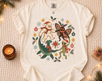 May include: An ivory t-shirt featuring a circular graphic of a yeti, a mothman, a loch ness monster, and an alien, all wearing red and white striped scarves and Santa hats, surrounded by Christmas ornaments, holly, and snowflakes.
