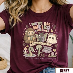 May include: Berry-colored t-shirt with a whimsical graphic featuring various cartoon creatures and the text "WE'RE ALL WEIRD SOMETIMES." The design includes an alien, a ghost, a moth, and a sasquatch. The phrase "WE COME IN PEACE" is also visible.