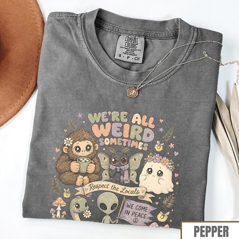May include: Pepper-colored t-shirt with the text "WE'RE ALL WEIRD SOMETIMES" and various cartoon characters. The shirt features a crew neck and short sleeves. A silver necklace with a pendant is draped across the shirt.