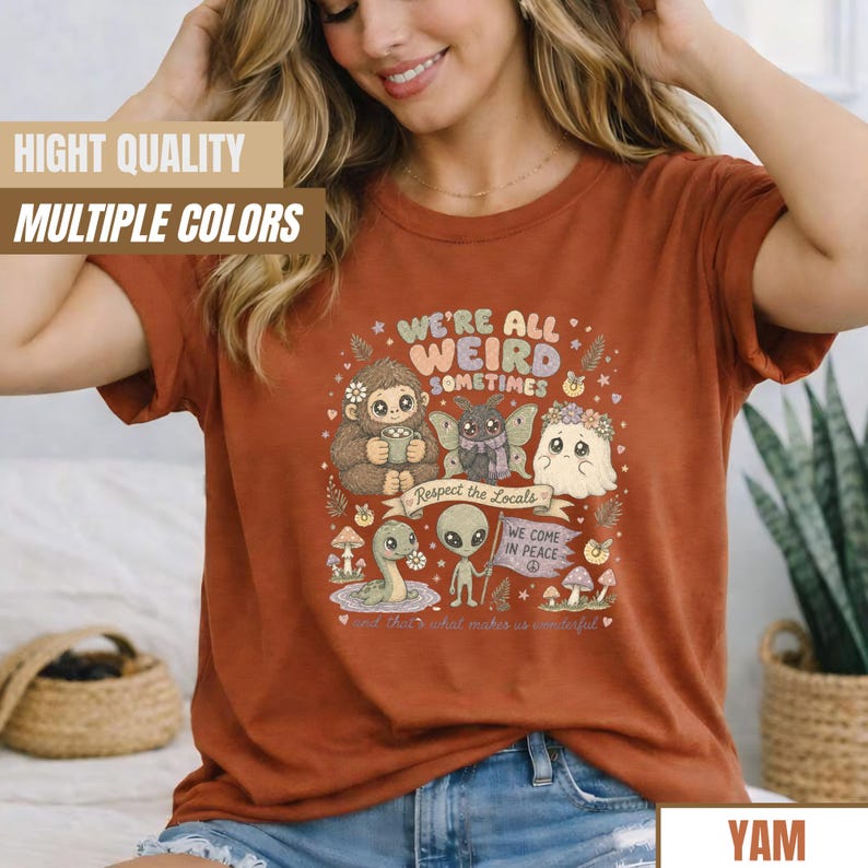 May include: Rust-colored t-shirt with the text "WE'RE ALL WEIRD SOMETIMES" and various cartoon characters. The shirt also features the phrases "Respect the Locals" and "WE COME IN PEACE".