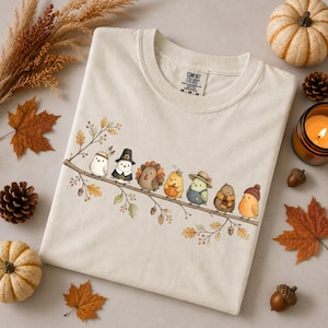 Peut inclure: Un t-shirt couleur crème avec un motif représentant sept petits oiseaux perchés sur une branche. Les oiseaux portent diverses tenues sur le thème de l'automne, notamment des chapeaux et des écharpes. Le t-shirt porte une étiquette Comfort Colors.