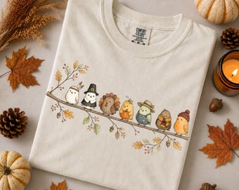 May include: A cream-colored t-shirt featuring a graphic of seven small birds perched on a branch. The birds are dressed in various autumn-themed outfits, including hats and scarves. The shirt has a Comfort Colors brand label at the neck.