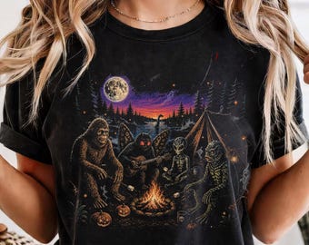 May include: A black t-shirt featuring a graphic print of Bigfoot, a moth-like creature, an alien, and a swamp creature sitting around a campfire at night. The scene includes a full moon, a tent, pine trees, and pumpkins under a starry sky.