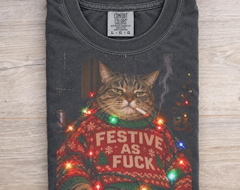 Könnte beinhalten: Ein anthrazitfarbenes T-Shirt mit einem Aufdruck einer getigerten Katze, die einen roten Weihnachtspullover mit dem Text "FESTIVE AS FUCK" trägt, eine Zigarette im Mund hat und in bunte Lichterketten gewickelt ist. Das T-Shirt hat ein Comfort Colors Etikett.
