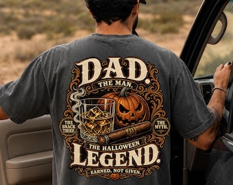 May include: A charcoal gray t-shirt featuring a back graphic with a glass of whiskey, a lit cigar, and a carved jack-o'-lantern. The text reads: DAD. THE MAN. THE SNACK THIEF. THE MYTH. THE HALLOWEEN LEGEND. EARNED, NOT GIVEN. The design uses orange and cream.