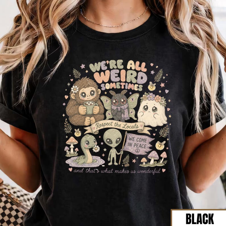 May include: Black graphic tee featuring a whimsical design with the text "WE'RE ALL WEIRD SOMETIMES." The design includes illustrations of a Bigfoot, a moth, a ghost, a snake, and an alien. The phrase "Respect the Locals" is also present.