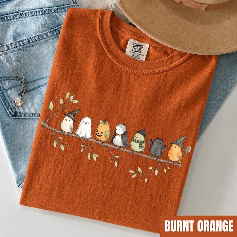 Peut inclure: Un t-shirt orange br&ucirc;l&eacute; avec un motif de huit petits oiseaux perch&eacute;s sur une branche, chacun portant un costume d'Halloween diff&eacute;rent, notamment une sorci&egrave;re, un fant&ocirc;me, une citrouille, un vampire et une chauve-souris. &Eacute;tiquette Comfort Colors.