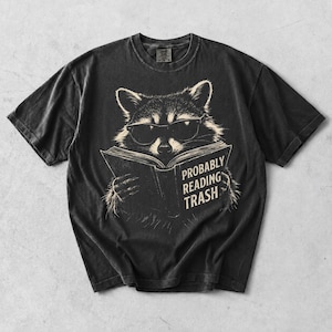 May include: A charcoal black short-sleeve t-shirt featuring a graphic illustration of a raccoon wearing sunglasses and reading an open book. The text on the book cover reads, "PROBABLY READING TRASH" in a bold, off-white, sans-serif typeface.