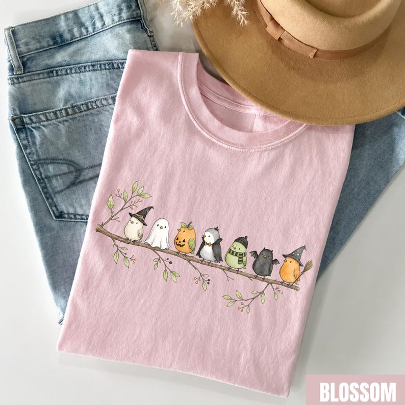 Peut inclure: Un t-shirt rose clair orn&eacute; d'un motif repr&eacute;sentant huit petits oiseaux perch&eacute;s sur une branche, chacun portant un costume d'Halloween diff&eacute;rent, notamment un chapeau de sorci&egrave;re, un drap de fant&ocirc;me, une citrouille, un smoking, une &eacute;charpe, des ailes de chauve-souris et un chapeau de magicien, avec un jean bleu et un chapeau beige.