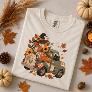 May include: A cream-colored t-shirt featuring a graphic of a green vintage pickup truck carrying pumpkins, sunflowers, and a black cat wearing a witch hat. The truck has plaid and floral patterns, and the shirt has a Comfort Colors brand label.