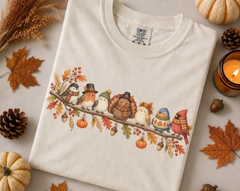 May include: A cream-colored t-shirt featuring a graphic of various birds perched on a branch. The birds are wearing autumn-themed accessories like hats and scarves. The graphic includes fall foliage, acorns, and small pumpkins. The shirt has a Comfort Colors label.