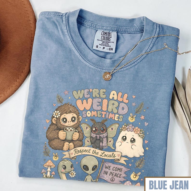 May include: Blue jean colored t-shirt with the text "WE'RE ALL WEIRD SOMETIMES" and a graphic of whimsical creatures. The shirt has a crew neck and short sleeves. A gold necklace with a pendant is on the shirt.