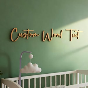 May include: A white wooden cot with a grey textured blanket draped over the front rail. Above the cot, the words Custom Wood Text are mounted on a green wall in a brown script font. A white cloud-shaped mobile hangs from a purple stand.