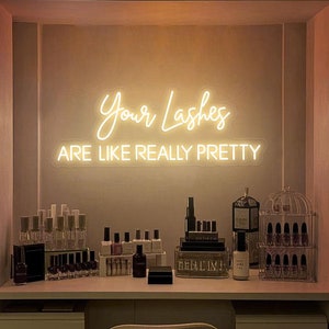 Könnte beinhalten: Ein Neon-Schild mit dem Text "Your Lashes Are Like Really Pretty" in einem weißen ovalen Rahmen. Das Schild leuchtet und hängt an einer Wand über einer Theke mit verschiedenen Flaschen und Gläsern von Schönheitsprodukten.