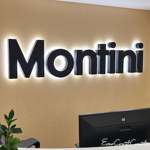 Könnte beinhalten: Schwarzes, beleuchtetes "Montini"-Schild an einer strukturierten, beigefarbenen Wand. Die Buchstaben sind fett und modern, mit hellem Licht, das jede Buchstaben umrandet. Ein Computermonitor und ein Schreibtisch sind im Vordergrund zu sehen.