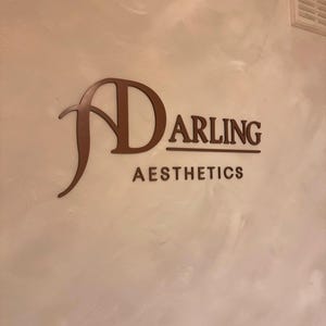K&ouml;nnte beinhalten: Ein Wandschild mit dem dunkelbraunen Schriftzug "DARLING AESTHETICS", wobei ein stilisiertes, &uuml;bergro&szlig;es "D" das Wort "ARLING" unterstreicht. Die Buchstaben sind auf einer hellen, strukturierten Wandoberfl&auml;che angebracht.