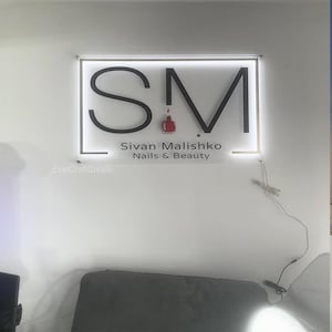 May include: A white wall with a black and white sign that says "SM Sivan Malishko Nails & Beauty" with a red nail polish bottle in the middle of the "M". The sign is lit up with white lights.