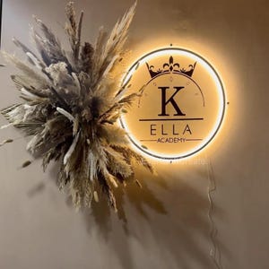 Può includere: Un'insegna a parete illuminata a forma circolare con la scritta "K ELLA ACADEMY" sotto una corona. A sinistra, una composizione decorativa di erbe e canne secche aggiunge un elemento naturale.