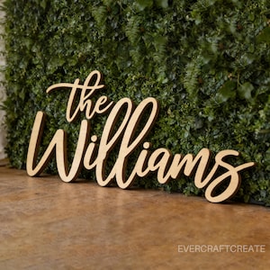 K&ouml;nnte beinhalten: Ein hellbraunes Holzschild mit der Aufschrift "The Williams" in einer kursiven Schriftart. Die Buchstaben sind als ein einziges St&uuml;ck ausgeschnitten und haben eine glatte, matte Oberfl&auml;che. Das Schild steht vor einer gr&uuml;nen Blattwand.