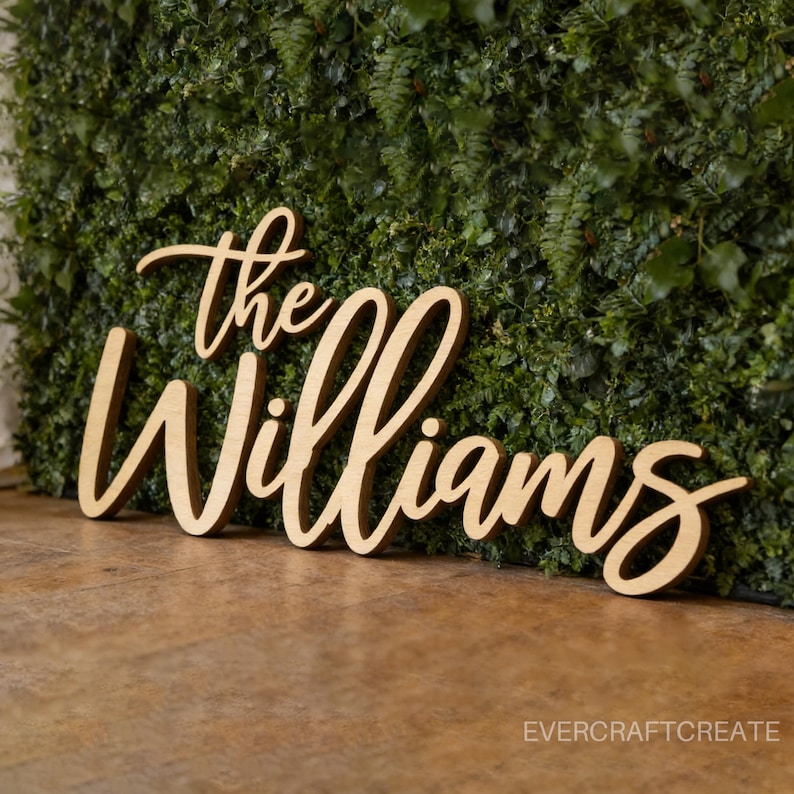 K&ouml;nnte beinhalten: Ein hellbraunes Holzschild mit der Aufschrift "The Williams" in einer kursiven Schriftart. Die Buchstaben sind als ein einziges St&uuml;ck ausgeschnitten und haben eine glatte, matte Oberfl&auml;che. Das Schild steht vor einer gr&uuml;nen Blattwand.