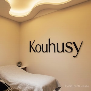 K&ouml;nnte beinhalten: Schwarze 3D-Buchstaben, die das Wort Kouhusy bilden, sind an einer schlichten beigen Wand angebracht. Unter dem Schriftzug steht eine Behandlungsliege, die mit einer grauen, strukturierten Decke bedeckt ist. Der Raum hat eine geschwungene Deckenleuchte.