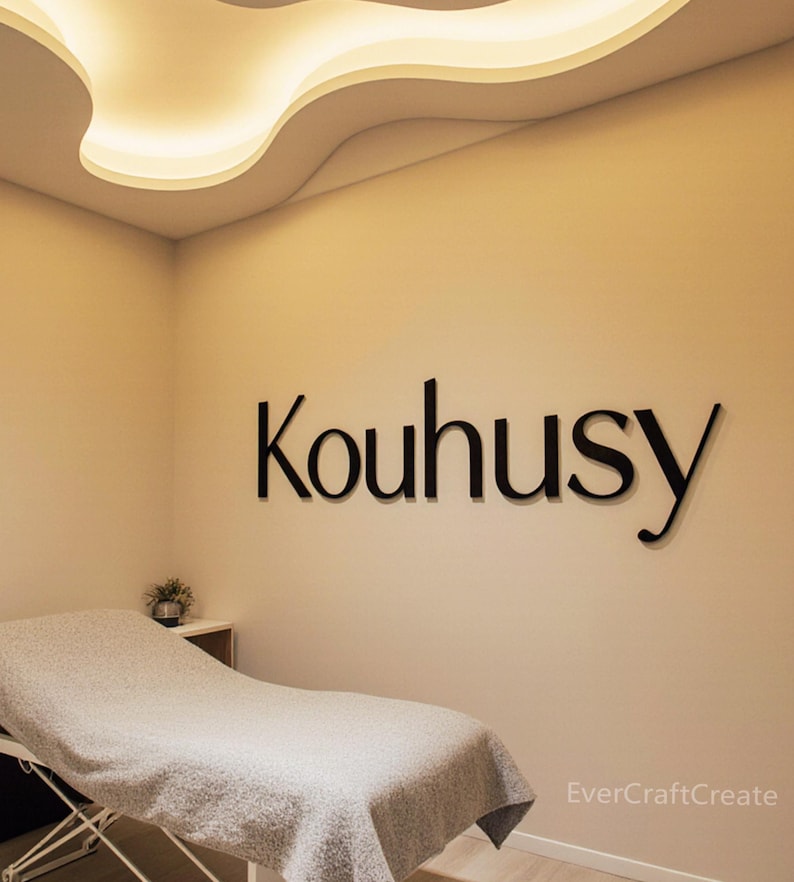 K&ouml;nnte beinhalten: Schwarze 3D-Buchstaben, die das Wort Kouhusy bilden, sind an einer schlichten beigen Wand angebracht. Unter dem Schriftzug steht eine Behandlungsliege, die mit einer grauen, strukturierten Decke bedeckt ist. Der Raum hat eine geschwungene Deckenleuchte.