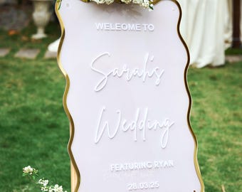 May include: White acrylic wedding sign with a gold border. The sign says "Welcome to Sarah's Wedding featuring Ryan" and the date "28.03.25".