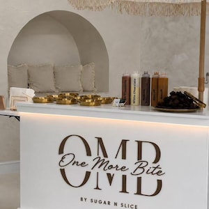 K&ouml;nnte beinhalten: Ein wei&szlig;er mobiler Catering-Wagen mit einem cremefarbenen Sonnenschirm mit Fransen. Die Vorderseite zeigt den braunen Schriftzug "OMB One More Bite BY SUGAR N SLICE". Auf der Theke stehen goldene Schalen, beschriftete Saucenflaschen und Brownies.