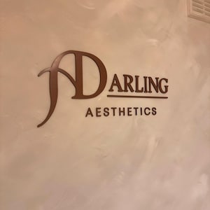 May include: A wall-mounted sign featuring the dark brown text "DARLING AESTHETICS" with a stylized, oversized "D" that curves around the word. The letters are mounted on a light-colored, textured wall surface with a subtle, swirled finish.