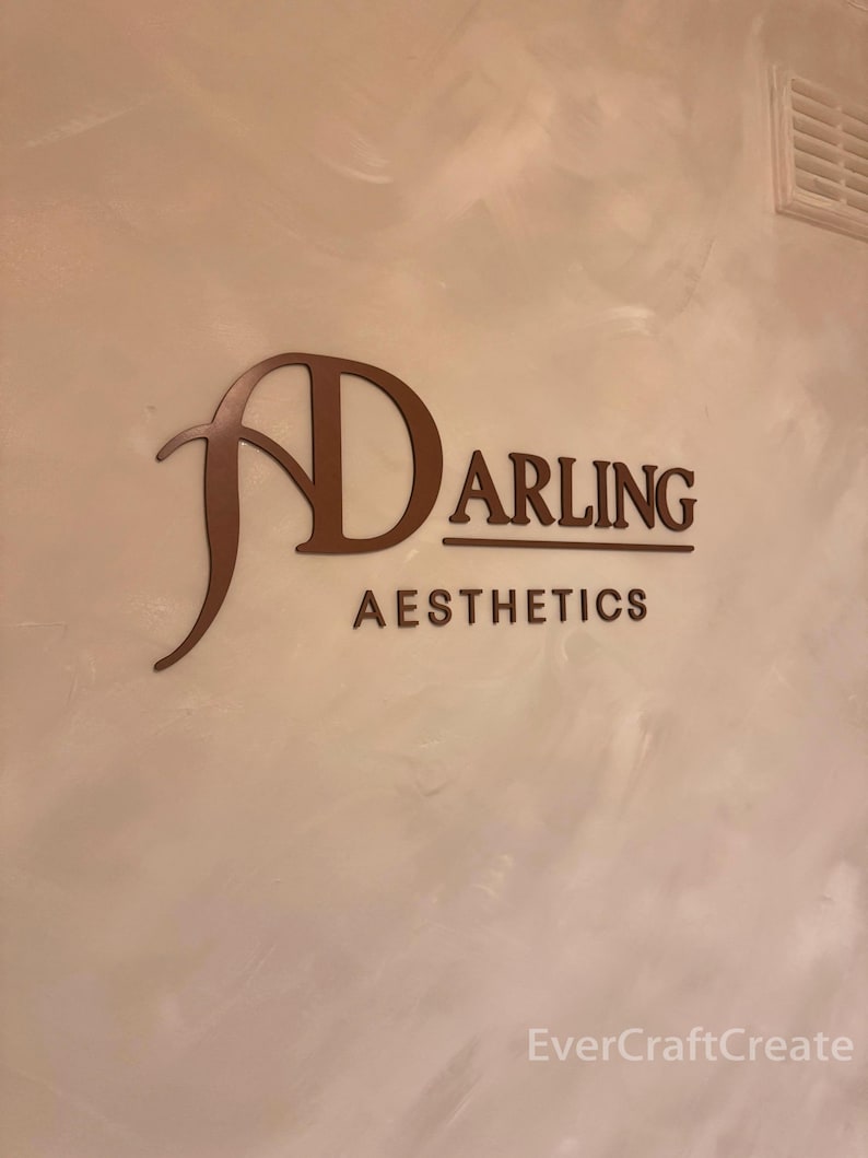 May include: A wall-mounted sign featuring the dark brown text "DARLING AESTHETICS" with a stylized, oversized "D" that curves around the word. The letters are mounted on a light-colored, textured wall surface with a subtle, swirled finish.