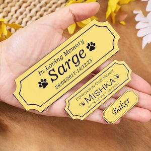 May include: Three yellow memorial plaques with black text and paw prints. The largest plaque reads "In Loving Memory Sarge 28/08/2017-14/12/23". The middle plaque says "FOREVER IN OUR HEART MISHKA 04/06/2006-01/06/2020". The smallest plaque says "Baker".