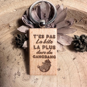 Peut inclure: Un porte-clés rectangulaire en bois avec un anneau argenté. Le porte-clés comporte du texte gravé et une image de dessin animé. Le texte se lit "T'ES PAS La bite LA PLUS dure du GANGBANG".
