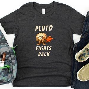 May include: A dark gray t-shirt with the text "PLUTO FIGHTS BACK" and a cartoon Pluto character. The image also includes a camouflage sling bag, blue jeans, and khaki slip-on shoes.
