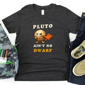 May include: A dark gray t-shirt with the text "PLUTO AIN'T NO DWARF" and a cartoon Pluto graphic, paired with blue jeans, khaki slip-on shoes, and a camouflage sling bag with markers and a ruler.