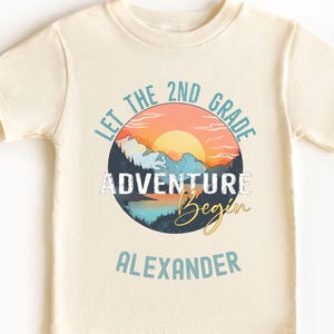 May include: Cream-coloured short-sleeved t-shirt with a circular graphic featuring a mountain landscape, sunset, and the text "LET THE 2ND GRADE ADVENTURE Begin ALEXANDER". The design incorporates blue, orange, and yellow hues.