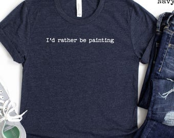 Puede incluir: Una camiseta azul marino con el texto "I'd rather be painting" en blanco. La camiseta se combina con unos vaqueros azules y zapatillas grises con cordones blancos. La camiseta está hecha de un material suave y cómodo.