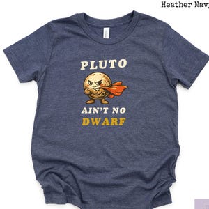 May include: Heather navy t-shirt with the text "PLUTO ain't no DWARF" in white and gold. The graphic features a cartoon Pluto with a red cape and angry expression. The shirt is a short-sleeved crew neck.