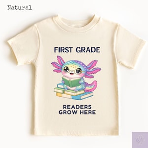May include: A natural-colored short-sleeve t-shirt featuring a graphic of a colorful axolotl wearing glasses and reading a book while sitting on a stack of books. The text FIRST GRADE and READERS GROW HERE is printed in dark blue above and below the graphic.