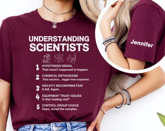 May include: Burgundy t-shirt with the text "UNDERSTANDING SCIENTISTS" and a list of science-related humorous statements. The sleeve has the name "Jennifer" printed on it. The shirt is paired with blue jeans.