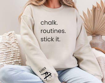May include: A light beige sweatshirt with the text "chalk. routines. stick it." printed on the front. The left sleeve has a black silhouette of a gymnast on a balance beam with the word "KAREN" above it.