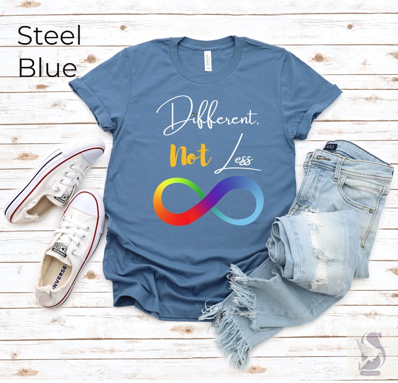 May include: Steel blue t-shirt featuring the text "Different, Not Less" and a rainbow infinity symbol. The image also shows light blue jeans and white trainers with red and blue detailing.
