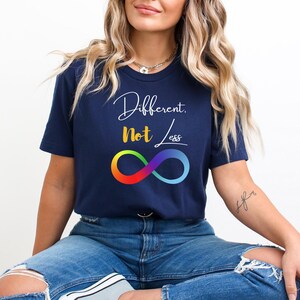 May include: A navy blue t-shirt featuring the text "Different, Not Less" in white script and yellow block letters. Below the text is a colourful infinity symbol. The t-shirt has a crew neck and is made of a soft material.
