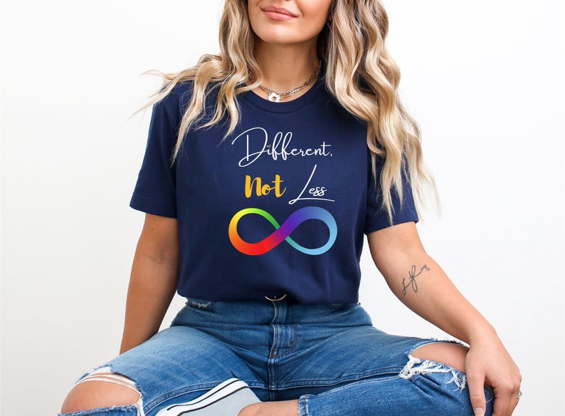 May include: A navy blue t-shirt featuring the text "Different, Not Less" in white script and yellow block letters. Below the text is a colourful infinity symbol. The t-shirt has a crew neck and is made of a soft material.
