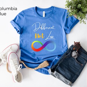 May include: A Columbia blue t-shirt with the words "Different, Not Less" and a rainbow infinity symbol. The shirt is paired with denim shorts, white trainers, and sunglasses. The shirt is a casual wear item.
