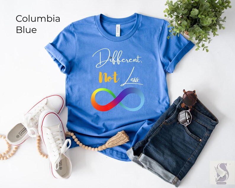 May include: A Columbia blue t-shirt with the words "Different, Not Less" and a rainbow infinity symbol. The shirt is paired with denim shorts, white trainers, and sunglasses. The shirt is a casual wear item.